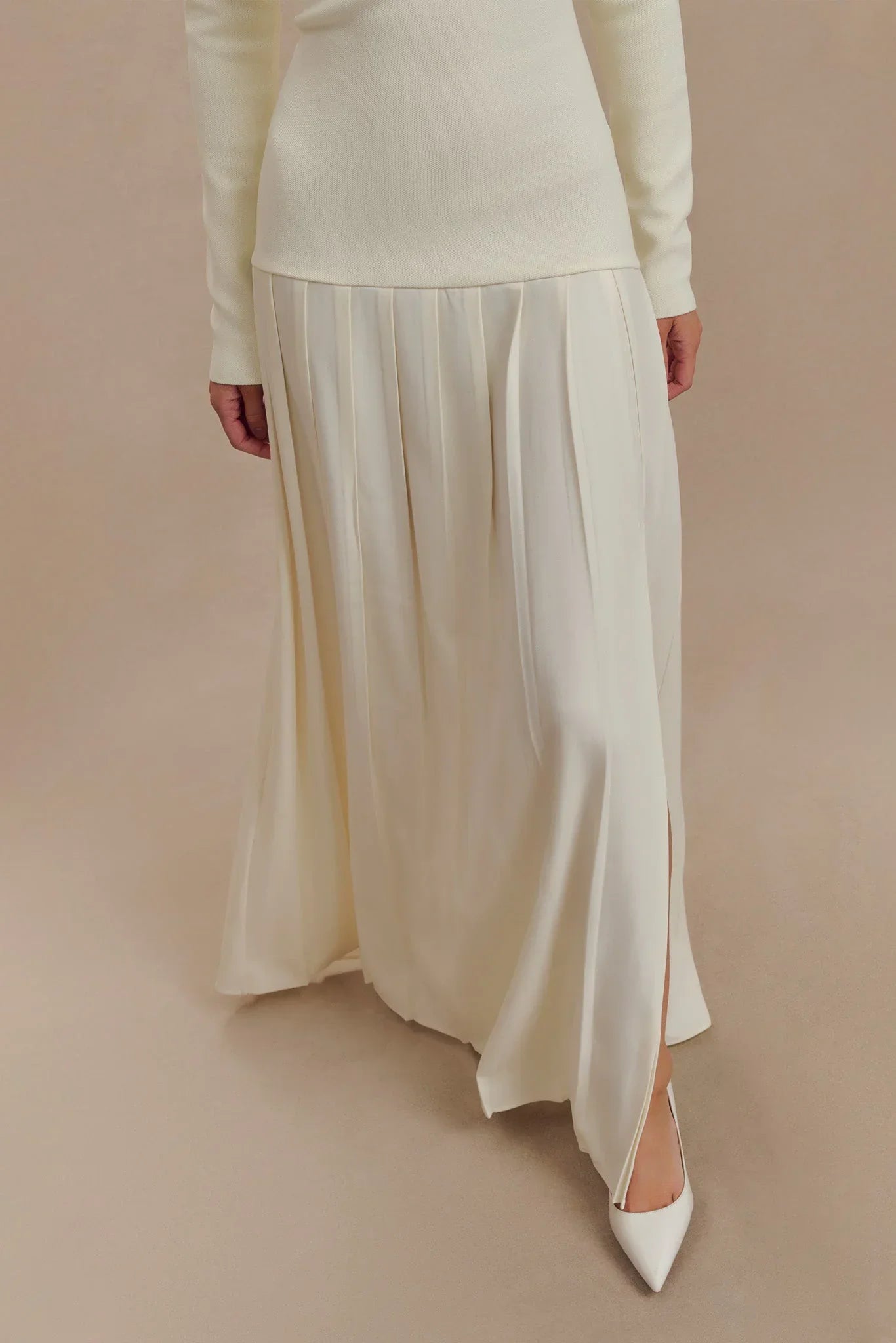A woman with long dark hair models a long-sleeved, off-white maxi dress featuring an off-the-shoulder ribbed bodice and a flowing pleated skirt with a side slit, standing against a solid beige background.