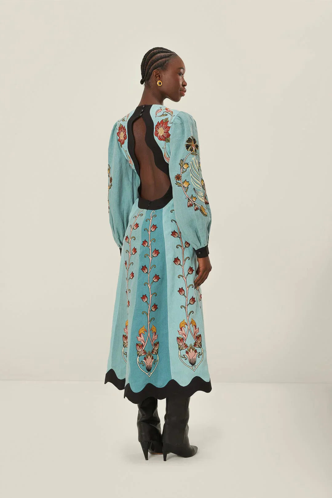 Full-length studio shot of a woman wearing a light blue, long-sleeved midi dress with a vibrant botanical print. The dress features a deep V-neckline, bishop sleeves, and contrasting black scalloped trim at the neck, waist, cuffs, and hem.