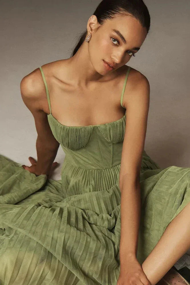 A woman models a sage green midi dress against a dark grey background. The dress has a structured bustier top with delicate spaghetti straps and a full, finely pleated skirt that falls to her lower calf.