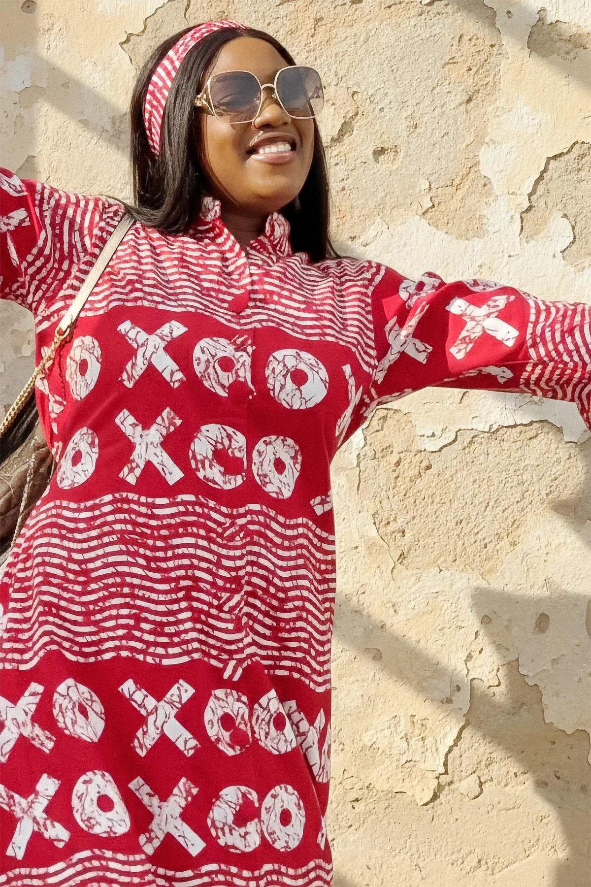 A woman wearing a red and white XOXO print maxi dress with a high neck, long flared sleeves, and a matching headband. She is smiling and standing with her arms out against a weathered beige wall.
