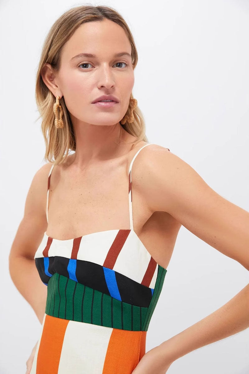 A full-length shot of a woman modeling a sleeveless midi dress with spaghetti straps. The dress has a straight, column silhouette and features a vibrant, multi-colored pattern of vertical stripes and color-blocked panels at the bodice and hem.