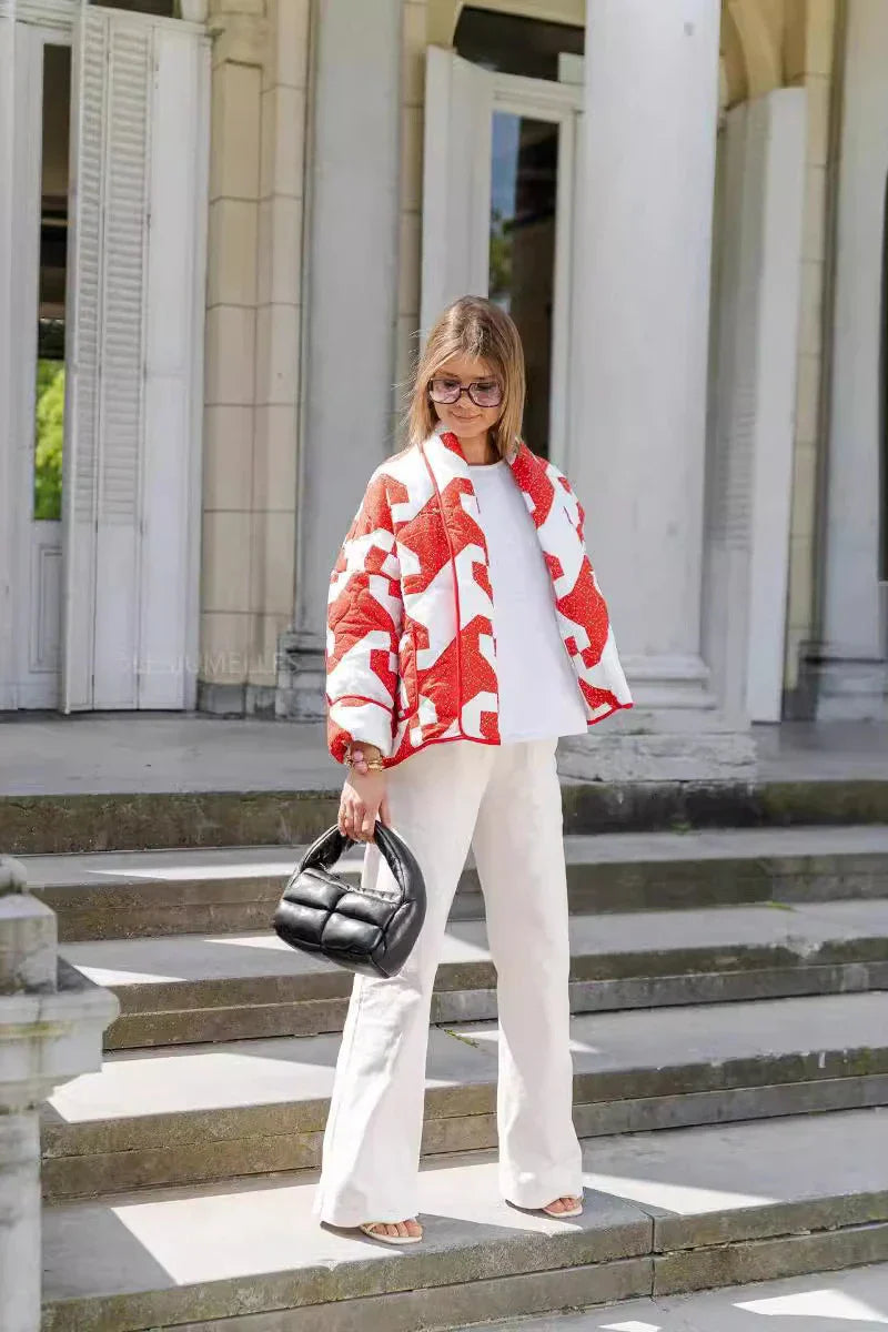 A woman with blonde hair and sunglasses wears a red and white quilted jacket with a geometric star pattern, styled over a white t-shirt and cream trousers, holding a black padded handbag.
