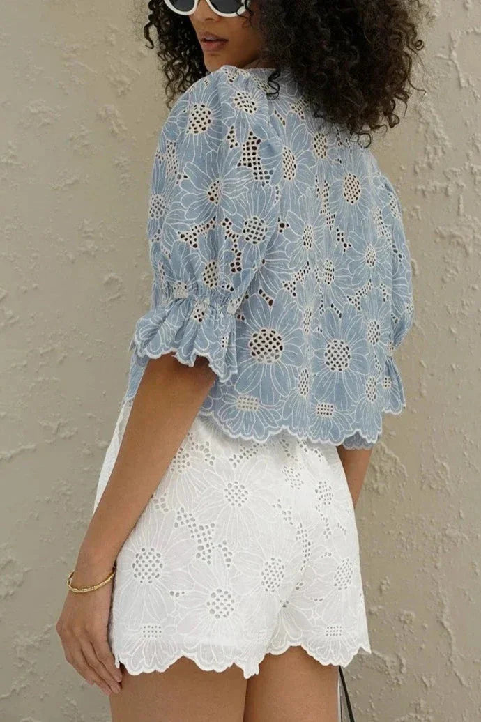 Woman in light blue floral eyelet blouse and white shorts holding straw bag.