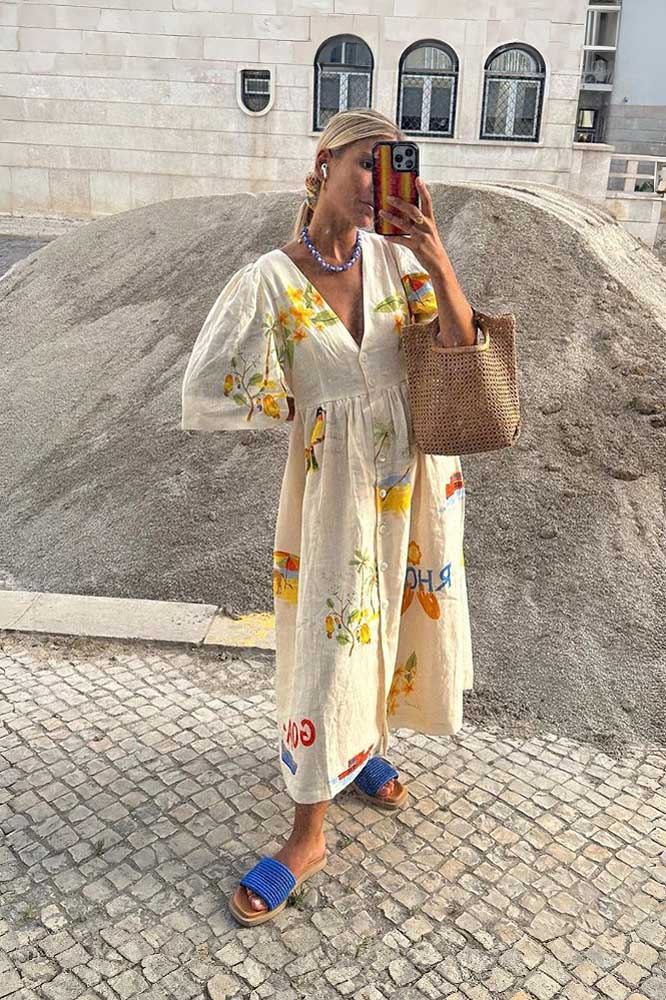 A woman wears a cream-colored, v-neck midi dress with elbow-length sleeves and a tropical vacation print. She holds a large brown woven tote bag and stands against a plain white background.