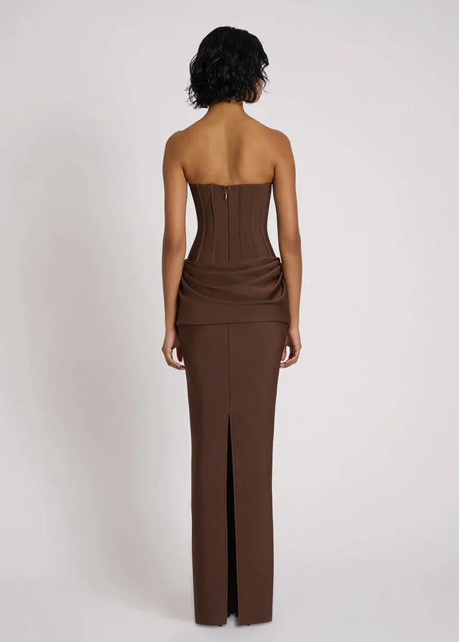 A woman wearing a formal, floor-length brown strapless dress featuring a structured corset bodice with vertical seaming and an elegant draped fabric detail at the hips.