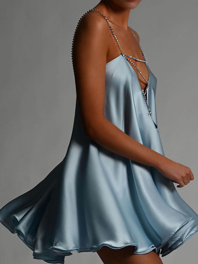 A woman wearing a chartreuse green satin mini dress with a plunging V-neckline created by a sparkling crystal chain harness that forms the straps and crosses over the chest. The dress has a loose, A-line silhouette and a ruffled hem, shown in motion against a grey background.