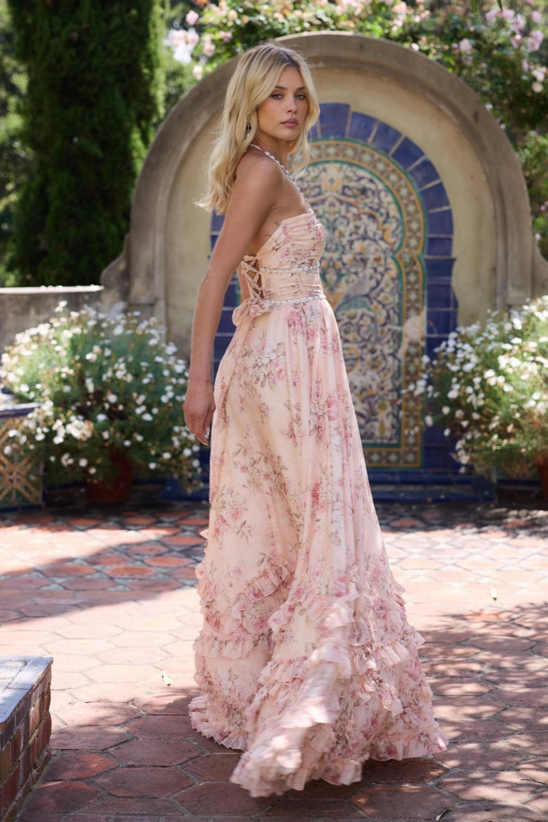 Pink - A woman posing in a light blue floral chiffon maxi dress with a beaded halter neckline, a high thigh-high slit, and a tiered ruffled hem, standing in front of a white garden gate.