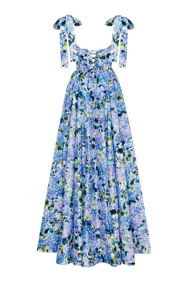 Full-length view of a model wearing an asymmetrical, tie-shoulder gown with a fitted bodice and a voluminous A-line skirt featuring a vibrant blue and purple hydrangea print, set against a white studio background.
