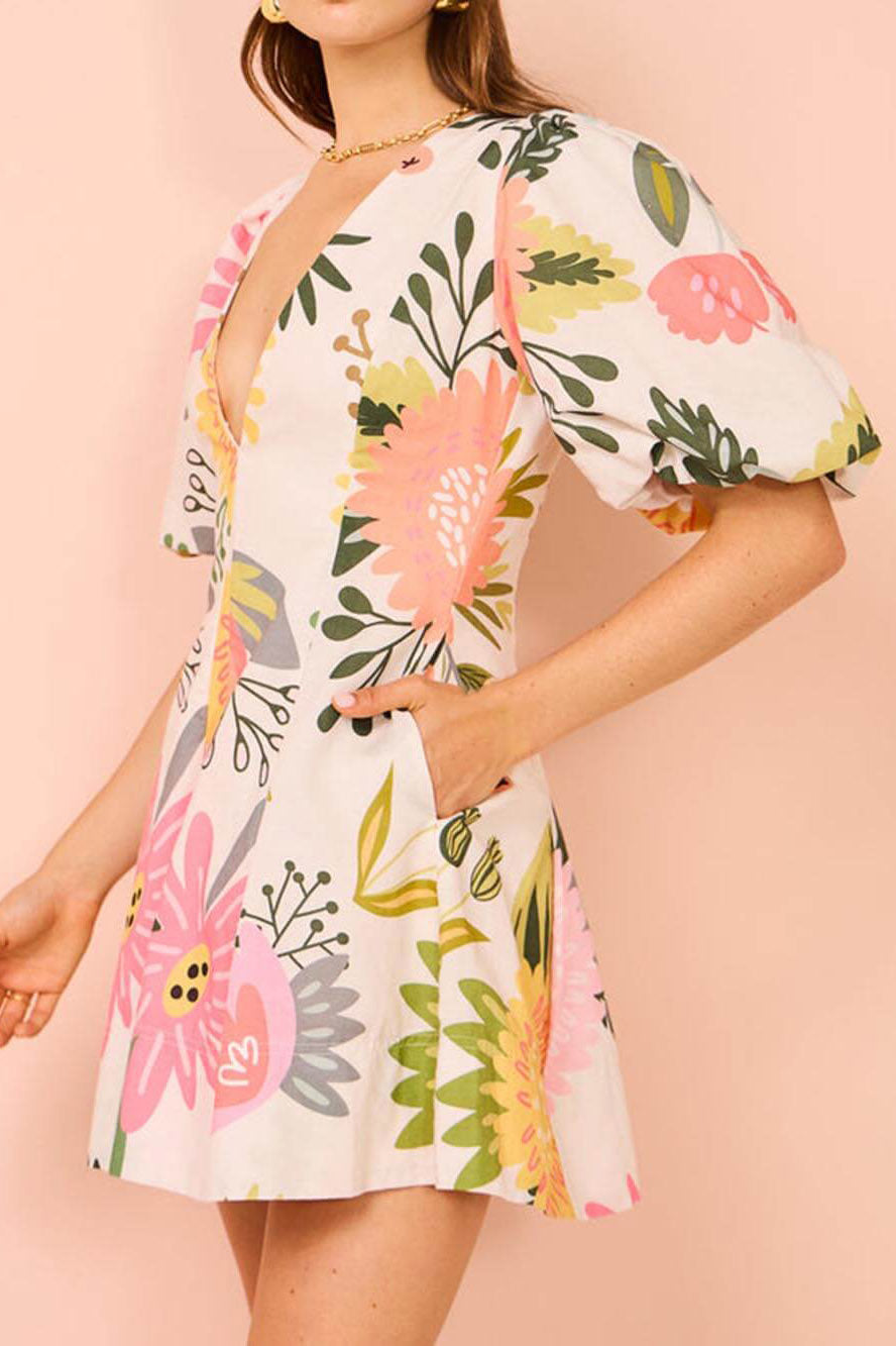 White - A woman wearing a white linen mini dress with a vibrant pink and yellow botanical print, featuring a deep V-neckline and voluminous puff sleeves.