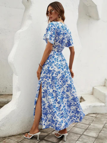 XL - A woman wearing a blue and white toile floral print chiffon maxi dress with flutter sleeves, a smocked waist, and a side slit, paired with a wide-brim straw hat and white strappy sandals.