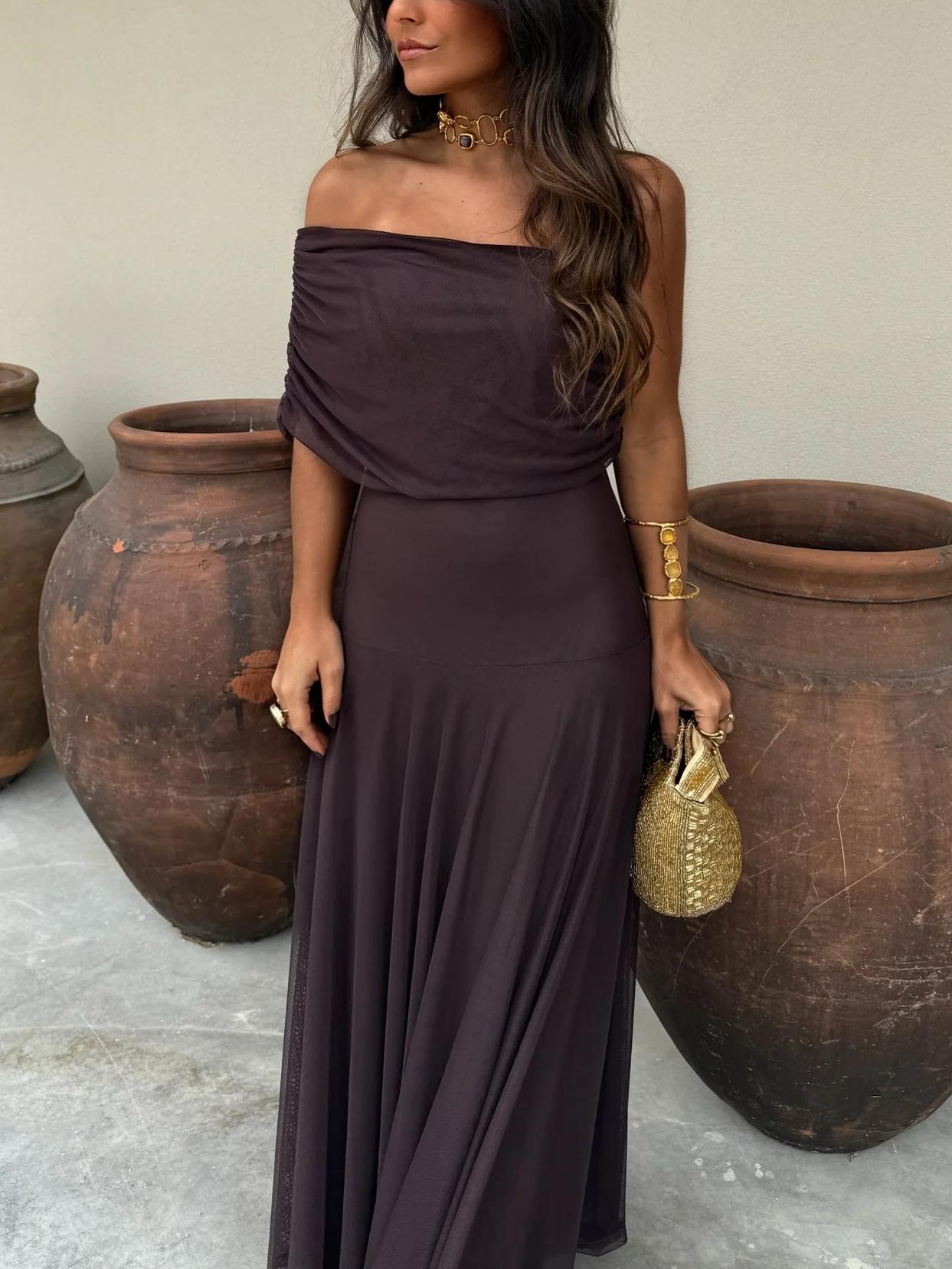 Brown - A woman wearing a dark brown, formal off-the-shoulder maxi dress featuring a ruched neckline and a flowing sheer mesh overlay over a fitted underlay, styled with gold accessories.