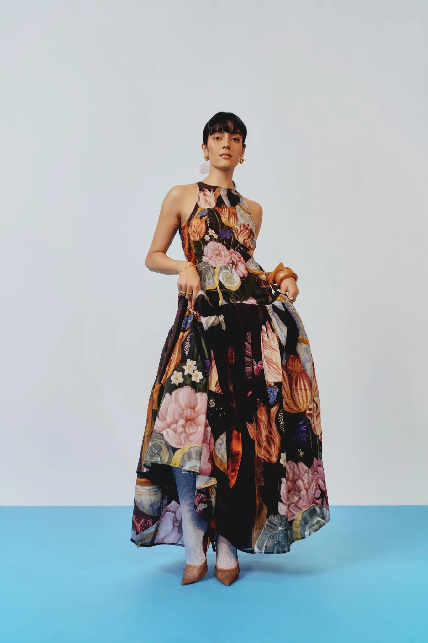 A woman with dark hair pulled back stands against a white background, wearing a sleeveless, high-neck maxi dress featuring a dark, painterly still-life print of flowers and vases.