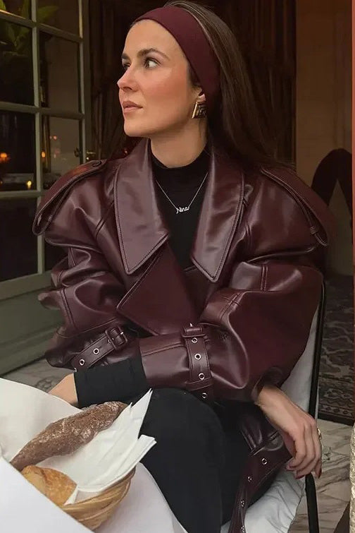 A woman wearing a burgundy oversized faux leather jacket with wide lapels, belted cuffs, and voluminous sleeves, styled over a black turtleneck in a city setting.
