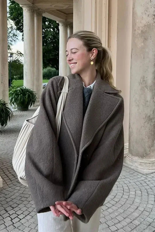 Grey - A woman wearing a taupe, short boxy jacket with oversized notch lapels, wide sleeves, and a double-breasted button front, styled over a black top.