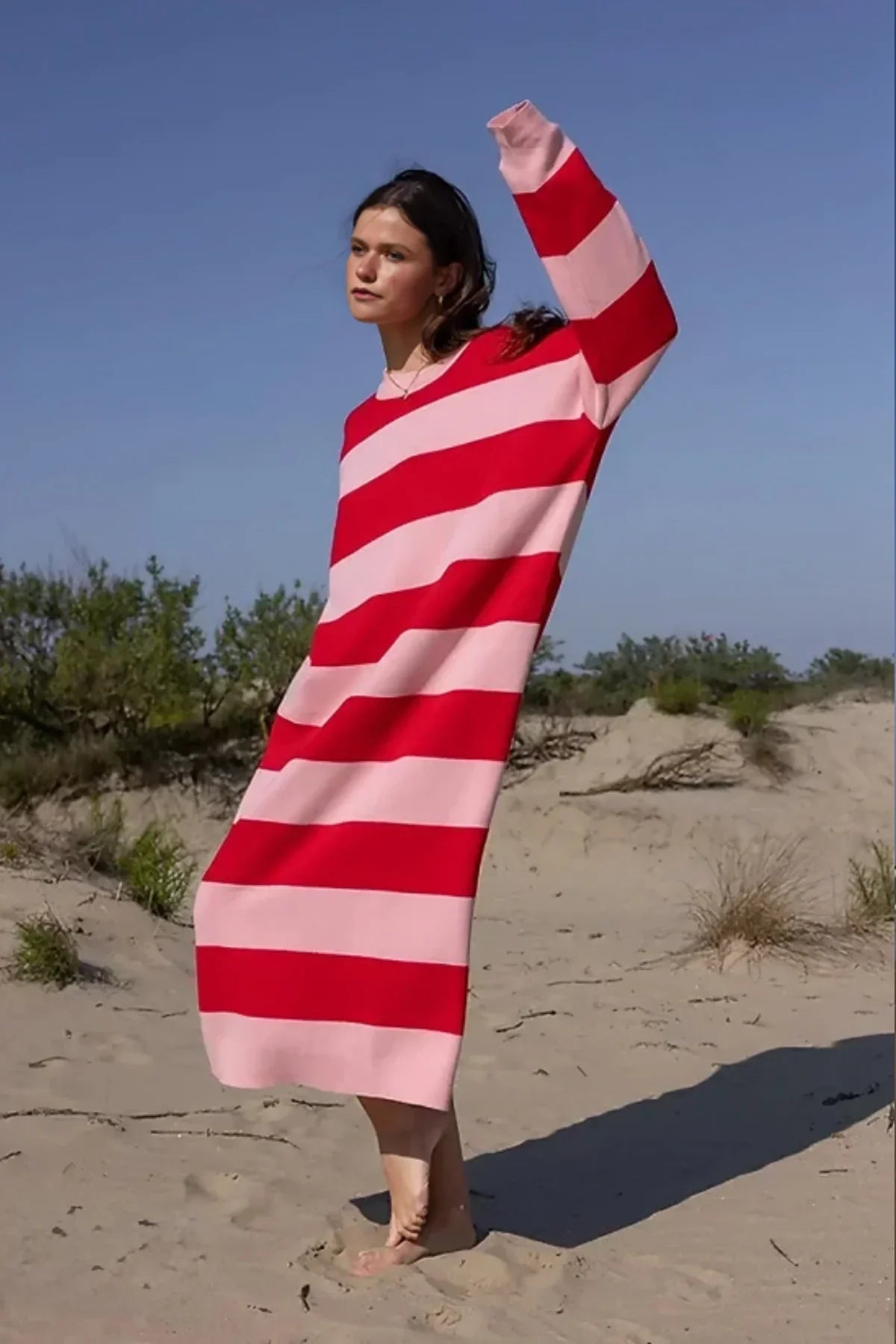 A woman standing on a sand dune, wearing a long-sleeved knit dress with bold red and pink horizontal stripes against a clear blue sky.