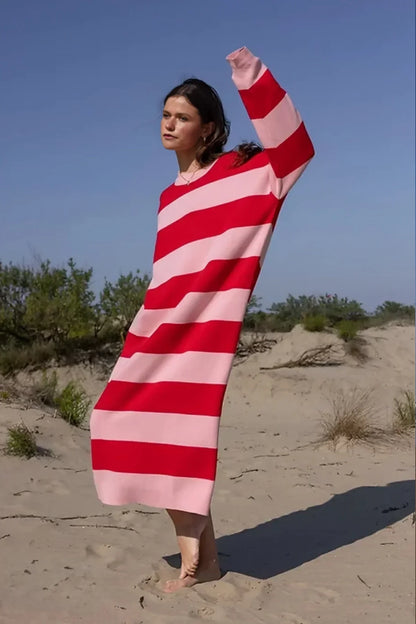 A woman standing on a sand dune, wearing a long-sleeved knit dress with bold red and pink horizontal stripes against a clear blue sky.