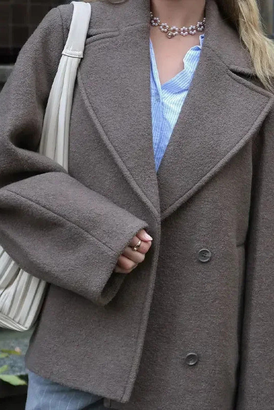 Grey - A woman wearing a taupe, short boxy jacket with oversized notch lapels, wide sleeves, and a double-breasted button front, styled over a black top.