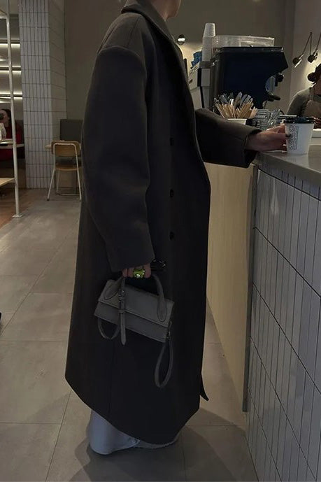 Brown - A woman wearing an oversized, dark brown double-breasted maxi coat with a notched lapel, styled over a beige sweater and light skirt.