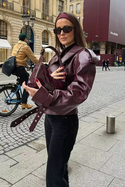 A woman wearing a burgundy oversized faux leather jacket with wide lapels, belted cuffs, and voluminous sleeves, styled over a black turtleneck in a city setting.