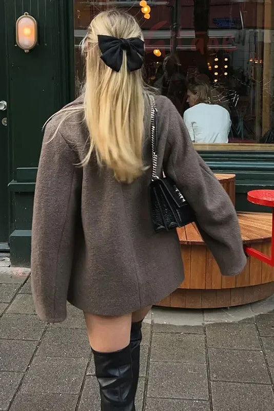 Grey - A woman wearing a taupe, short boxy jacket with oversized notch lapels, wide sleeves, and a double-breasted button front, styled over a black top.