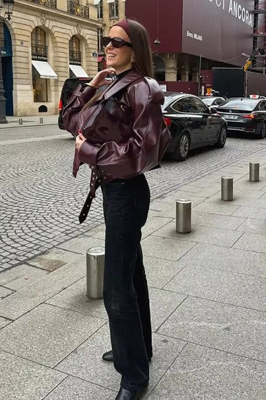 A woman wearing a burgundy oversized faux leather jacket with wide lapels, belted cuffs, and voluminous sleeves, styled over a black turtleneck in a city setting.