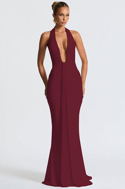 Red - A woman models a form-fitting, deep red halterneck maxi dress with a plunging neckline and extensive ruching, standing elegantly in a formal garden setting.