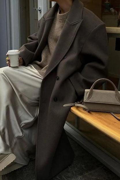 Brown - A woman wearing an oversized, dark brown double-breasted maxi coat with a notched lapel, styled over a beige sweater and light skirt.