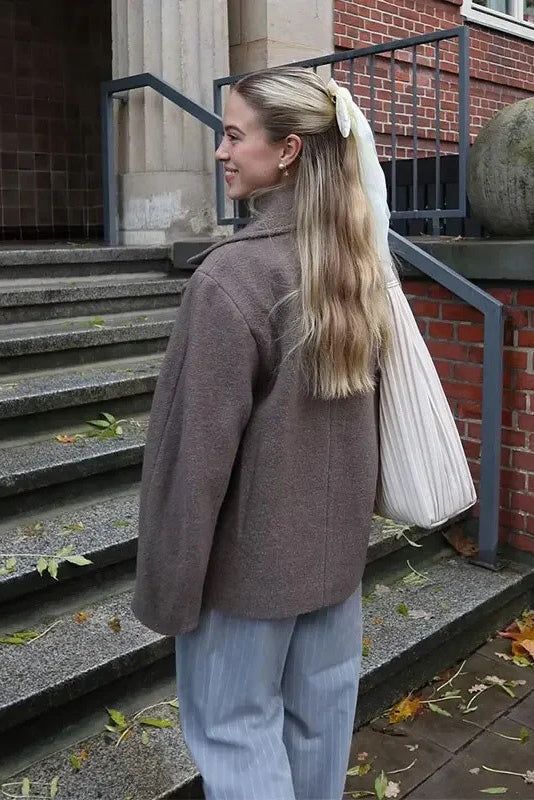 Grey - A woman wearing a taupe, short boxy jacket with oversized notch lapels, wide sleeves, and a double-breasted button front, styled over a black top.
