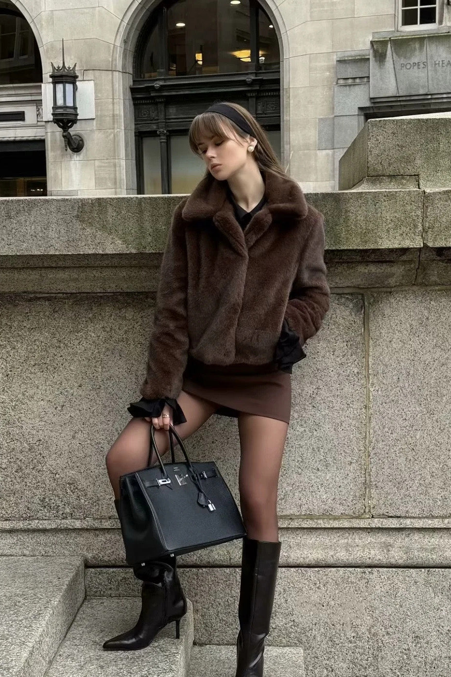 A woman standing outdoors on stone steps wearing a dark brown, plush faux fur cropped jacket with a wide collar, styled with a mini skirt and tall black boots.
