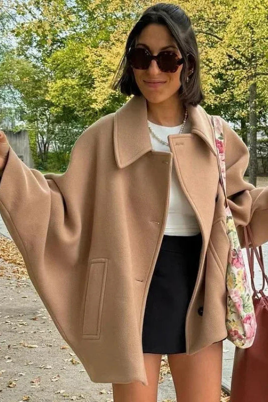 Black - A woman wearing a black oversized short coat with drop shoulders and a notched lapel, styled over a grey turtleneck and black shorts for a chic city look.
