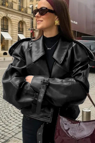 A woman wearing a burgundy oversized faux leather jacket with wide lapels, belted cuffs, and voluminous sleeves, styled over a black turtleneck in a city setting.