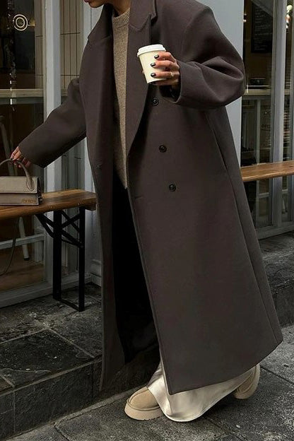 Brown - A woman wearing an oversized, dark brown double-breasted maxi coat with a notched lapel, styled over a beige sweater and light skirt.