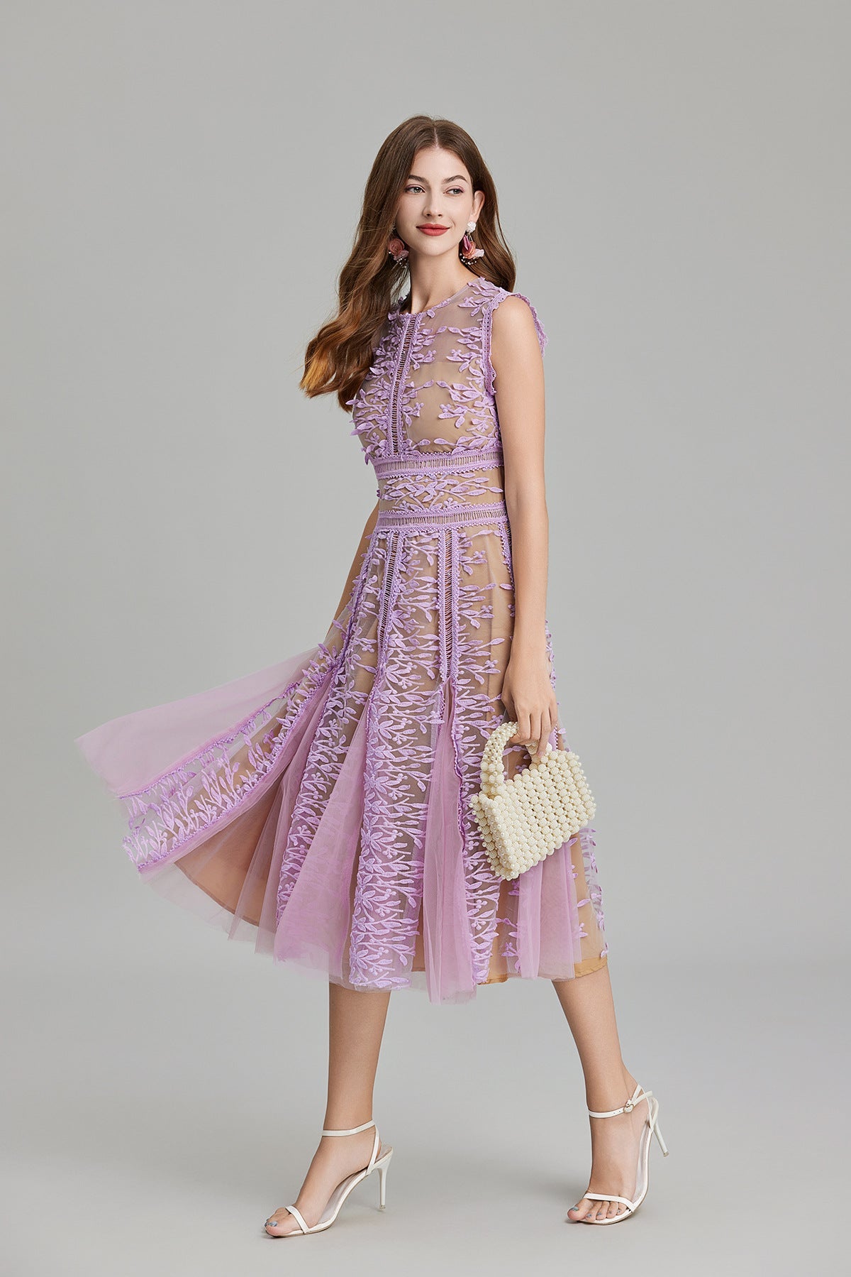 Full-length view of a woman wearing a sleeveless, sheer midi dress in pink and nude, featuring elaborate pink botanical embroidery in vertical panels and a flared tulle skirt.