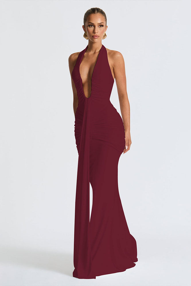 Red - A woman models a form-fitting, deep red halterneck maxi dress with a plunging neckline and extensive ruching, standing elegantly in a formal garden setting.