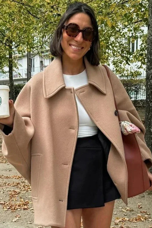 Black - A woman wearing a black oversized short coat with drop shoulders and a notched lapel, styled over a grey turtleneck and black shorts for a chic city look.