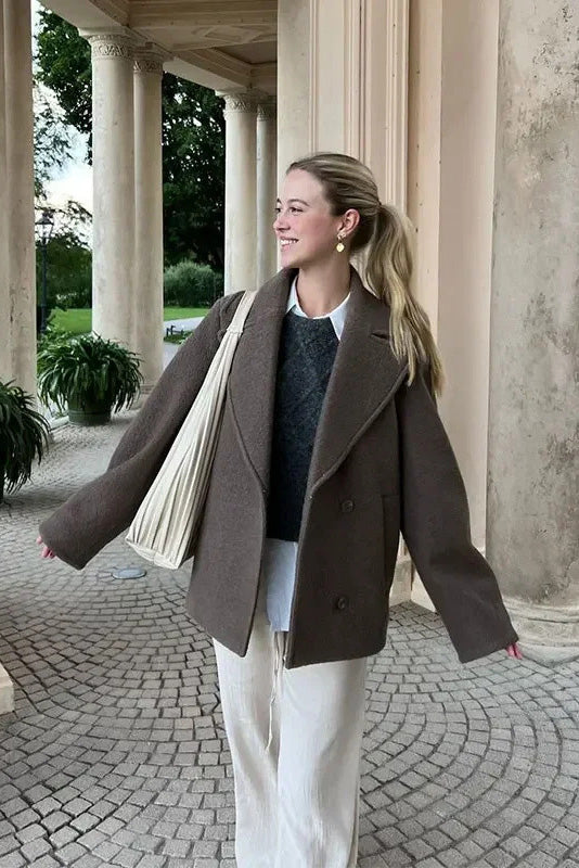 Grey - A woman wearing a taupe, short boxy jacket with oversized notch lapels, wide sleeves, and a double-breasted button front, styled over a black top.
