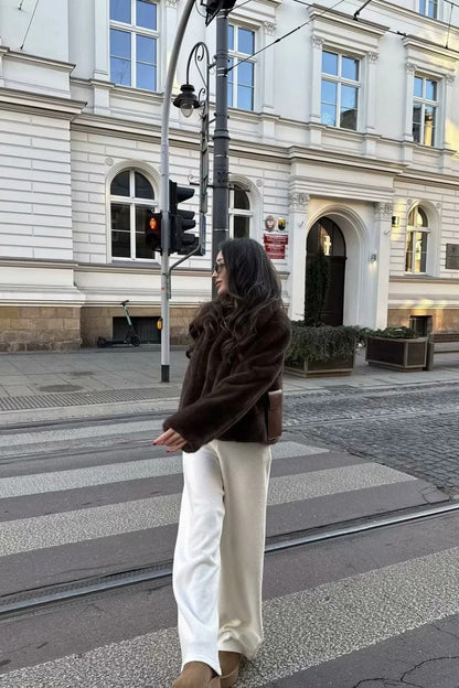 A woman standing outdoors on stone steps wearing a dark brown, plush faux fur cropped jacket with a wide collar, styled with a mini skirt and tall black boots.