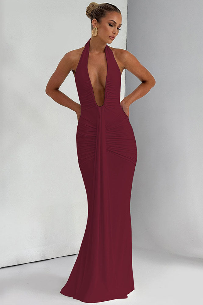 Red - A woman models a form-fitting, deep red halterneck maxi dress with a plunging neckline and extensive ruching, standing elegantly in a formal garden setting.