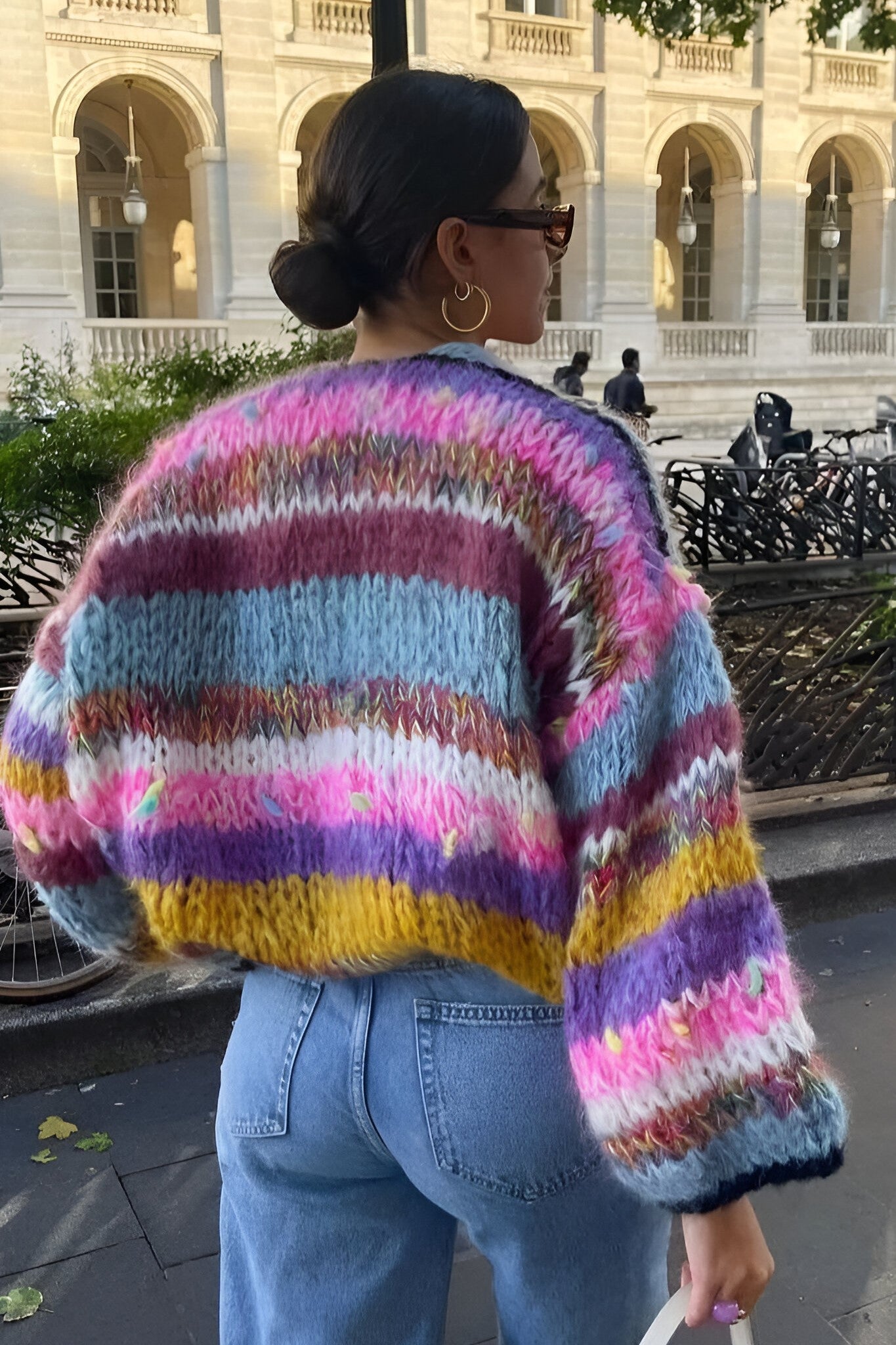 Rainbow - Woman wearing a vibrant, multicoloured striped chunky knit cropped cardigan layered over a white top, standing outdoors.