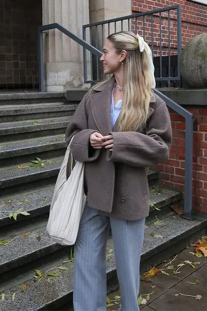 Grey - A woman wearing a taupe, short boxy jacket with oversized notch lapels, wide sleeves, and a double-breasted button front, styled over a black top.