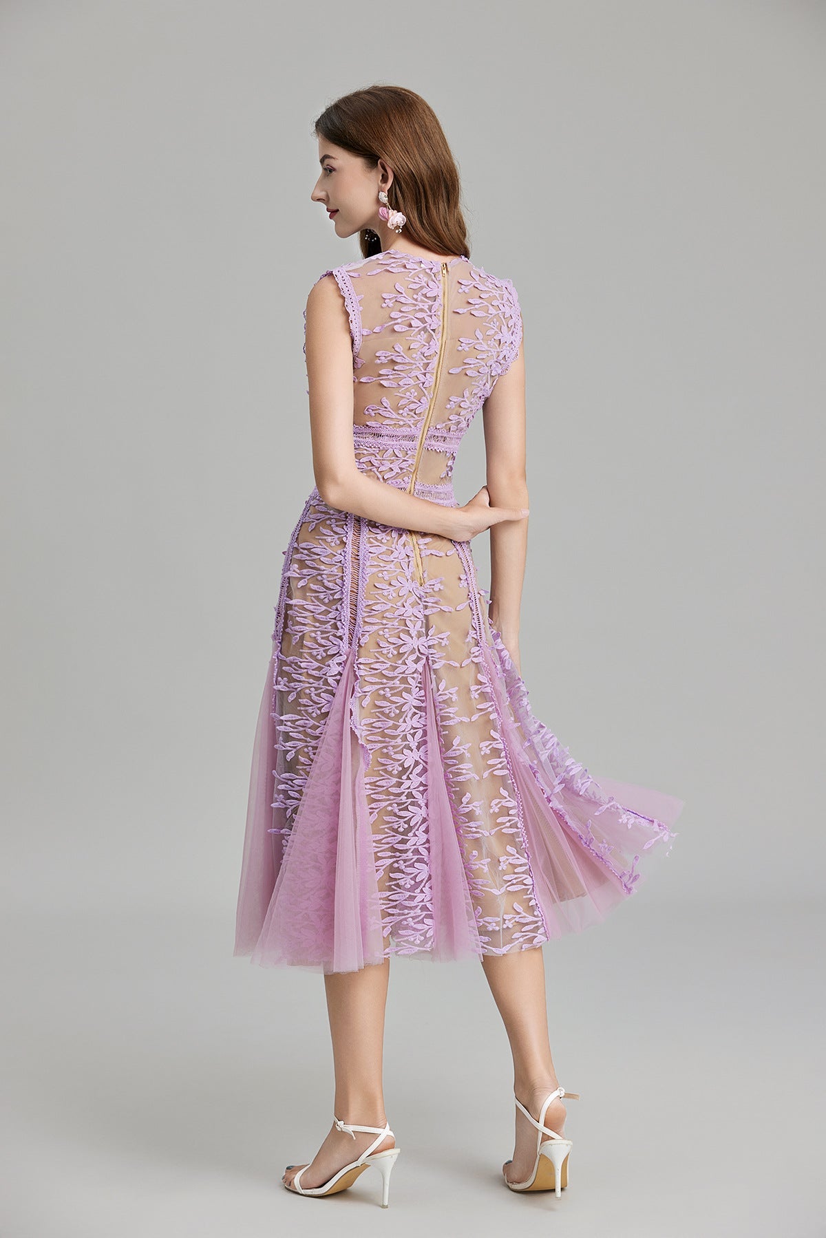 Full-length view of a woman wearing a sleeveless, sheer midi dress in pink and nude, featuring elaborate pink botanical embroidery in vertical panels and a flared tulle skirt.
