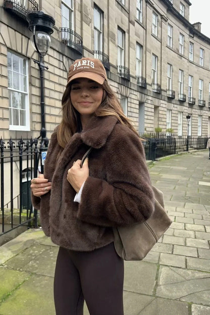 A woman standing outdoors on stone steps wearing a dark brown, plush faux fur cropped jacket with a wide collar, styled with a mini skirt and tall black boots.