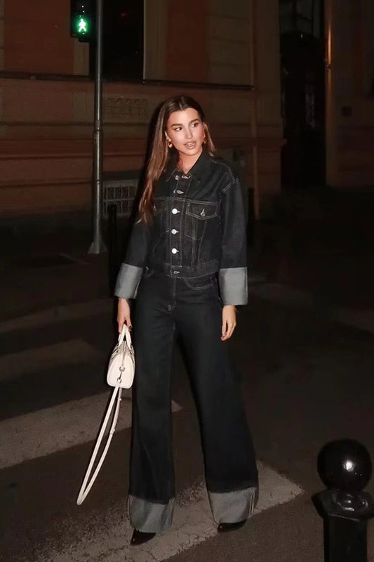 Woman wearing a dark, structured two-piece outfit consisting of a cropped jacket and high-waisted wide-leg trousers, both featuring prominent contrast turn-up cuffs, walking on a city street at night.