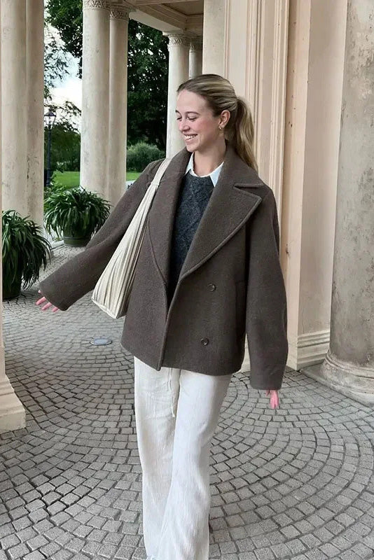 Grey - A woman wearing a taupe, short boxy jacket with oversized notch lapels, wide sleeves, and a double-breasted button front, styled over a black top.