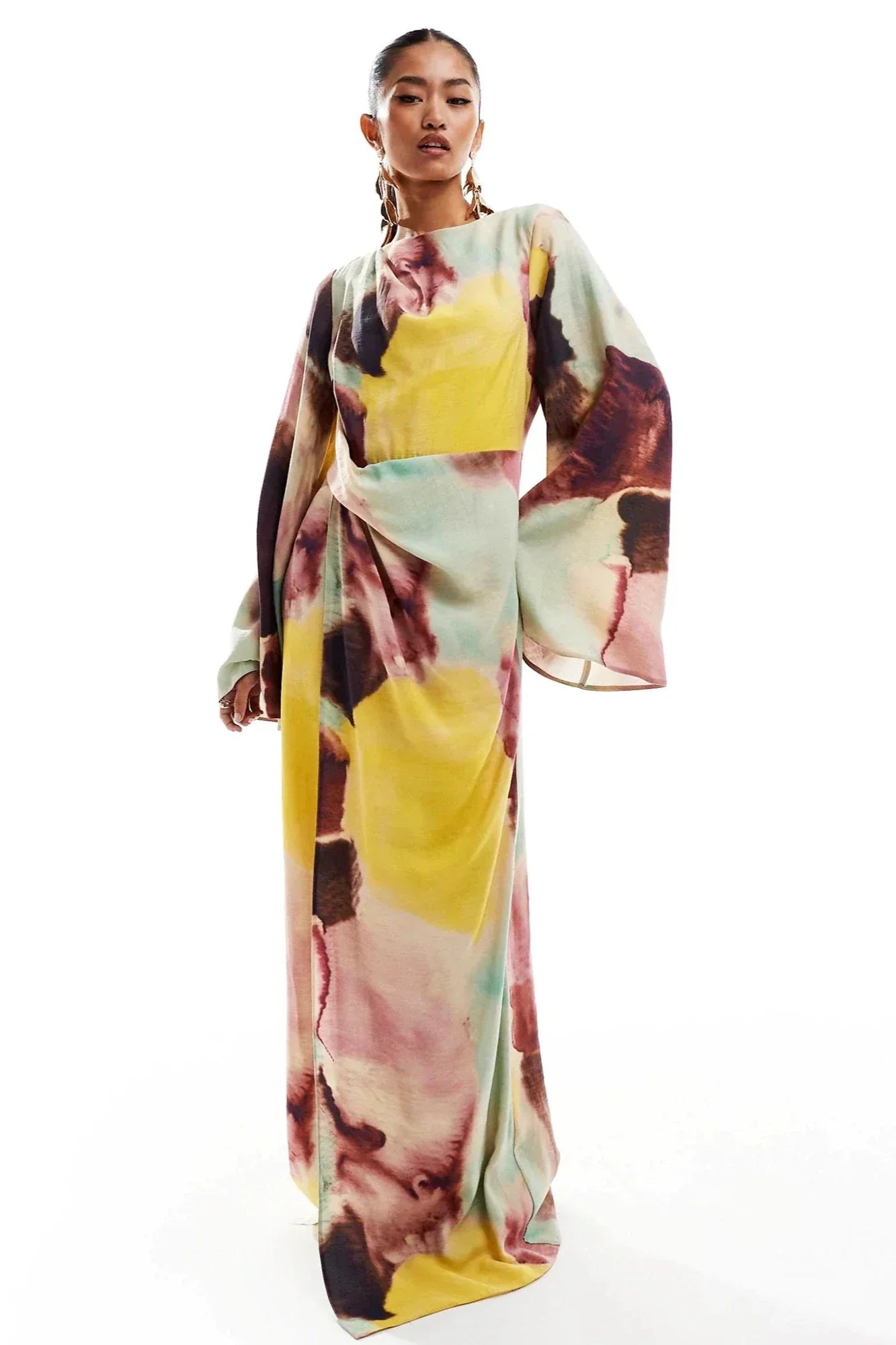 Watercolor - A woman models a long-sleeved maxi dress featuring a high neckline and a vibrant abstract watercolour print in yellow, pink, and brown. The dress has a twist-front detail at the waist and dramatic flared sleeves.