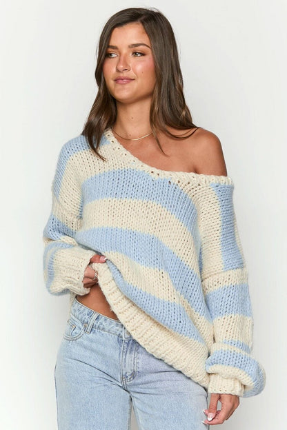 Blue - Woman wearing a chunky knit, slouchy off-shoulder jumper featuring wide horizontal stripes, paired with light wash denim jeans.