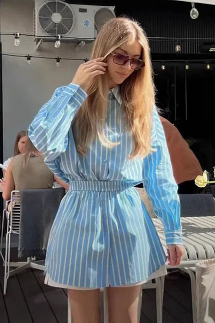 A woman wearing a blue and pale yellow vertically striped mini shirt dress featuring a collared neckline, button-down front, long sleeves, and a thick elasticated waistband.