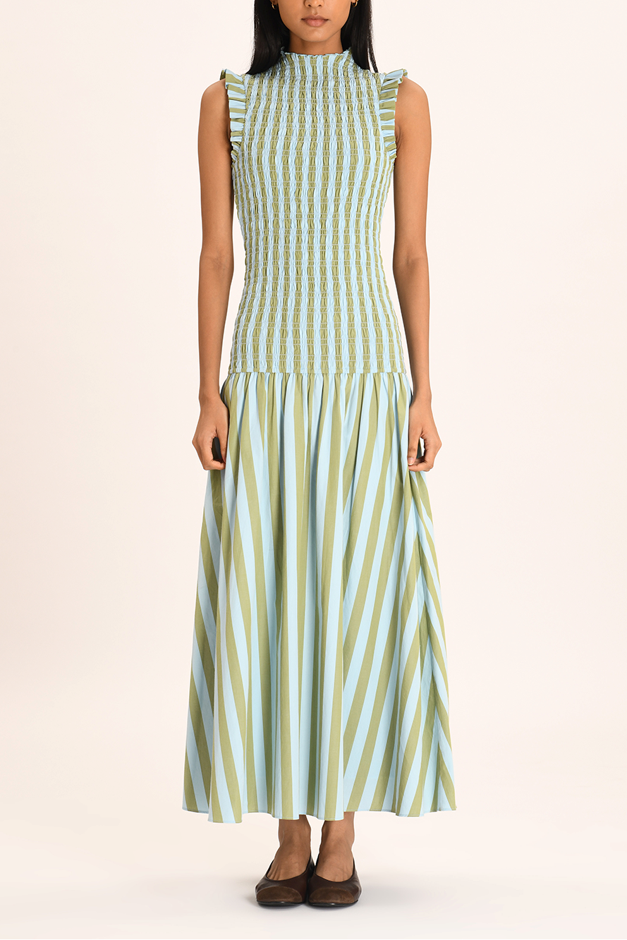 A woman models a sleeveless maxi dress with a high neckline and a fully smocked bodice. The dress features a pattern of vertical light blue and olive green stripes, ruffled shoulder details, and a full A-line skirt.