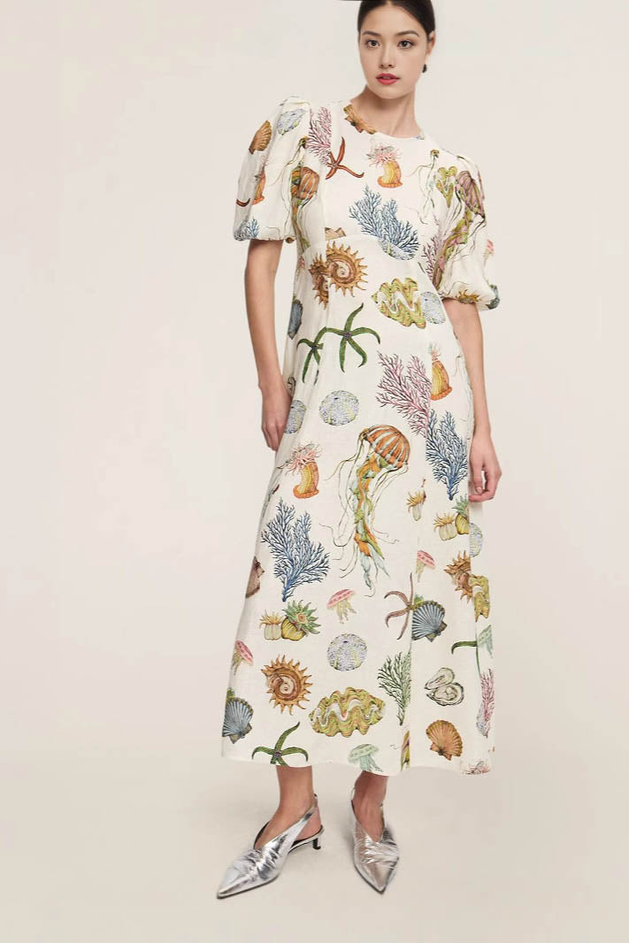 A woman models a short-sleeve, crewneck midi dress with a white background and a colorful, illustrative print of marine life including jellyfish, coral, and seashells.