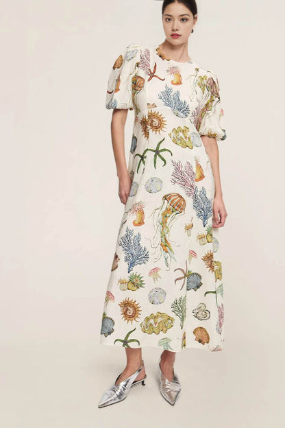 A woman models a short-sleeve, crewneck midi dress with a white background and a colorful, illustrative print of marine life including jellyfish, coral, and seashells.